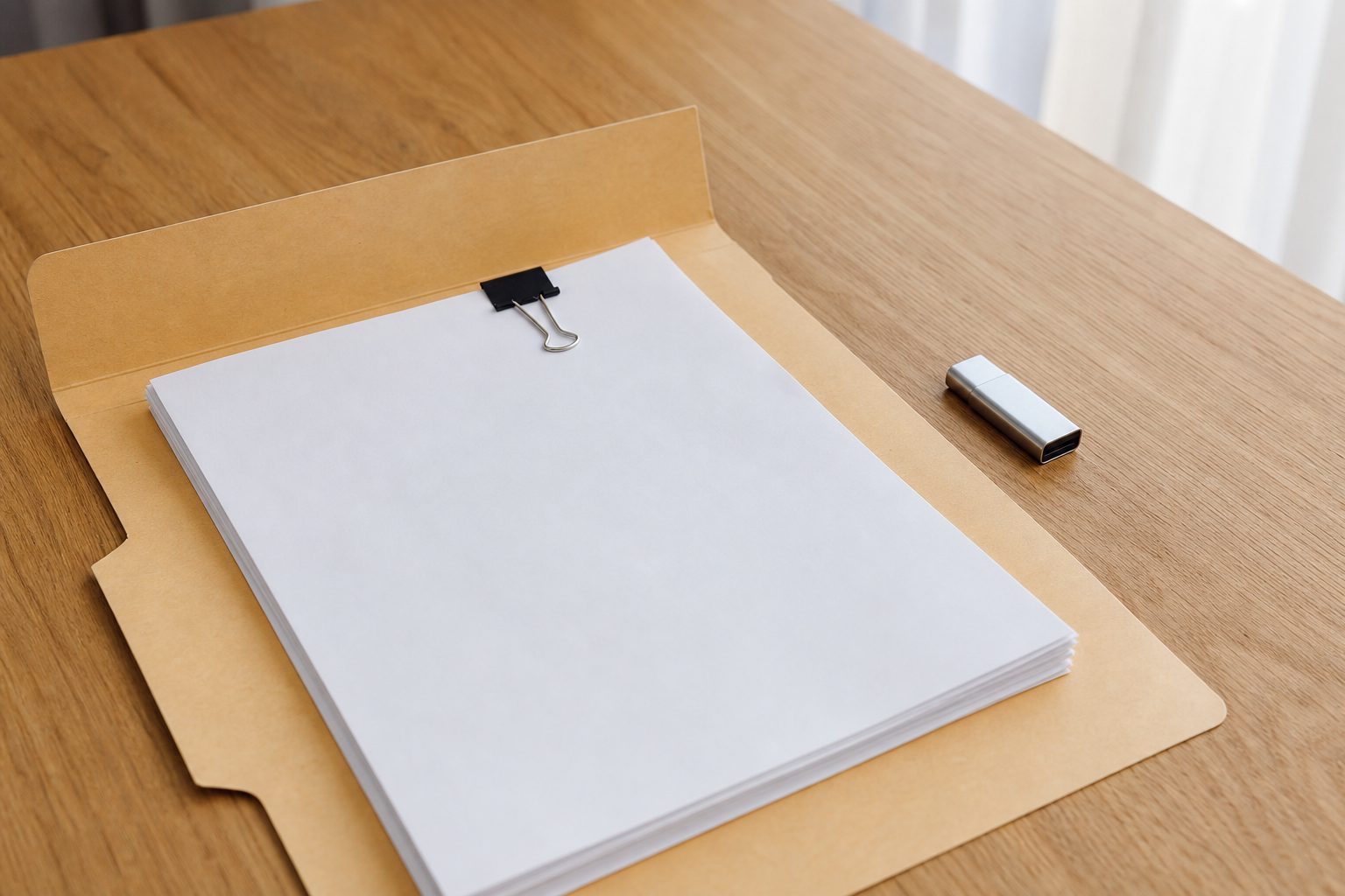 Open manila folder holding a stack of plain white paper held by a black binder clip, beside a small silver USB flash drive on an oak table