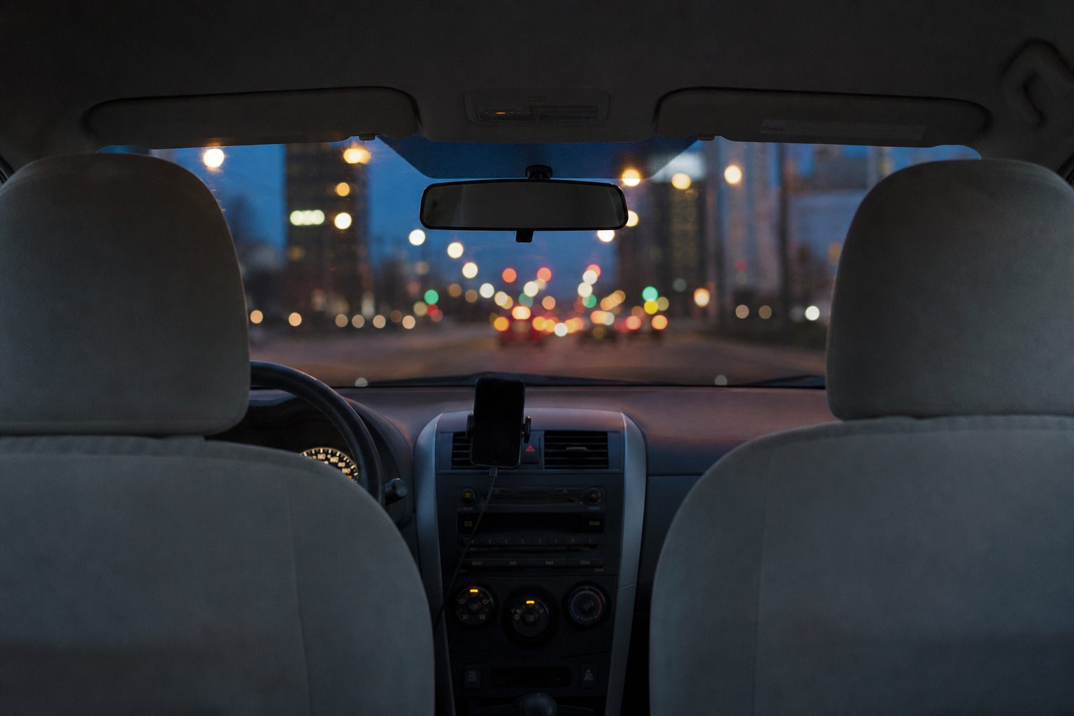 View from the back seat of a car at dusk, looking between two empty front seats toward a city street with blurred street lights